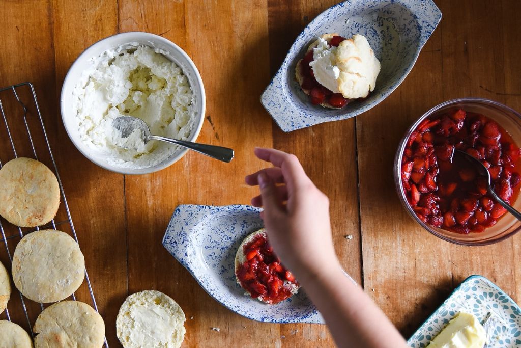 a finished strawberry shortcake with the ingredients around it and someone reaching for homemade whipped cream to add it to another one