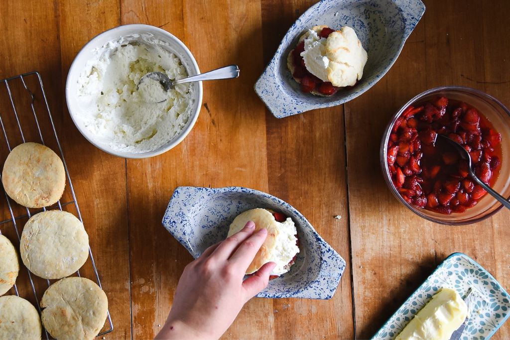 an finished  strawberry shortcake with the ingredients around it and someone placing the top on another one