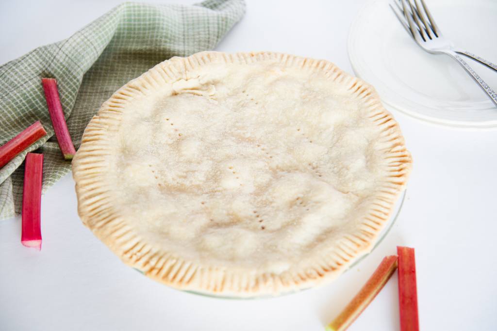 a pie sitting on a counter top with rhubarb stalks and a stack of plates with forks on it, next to it