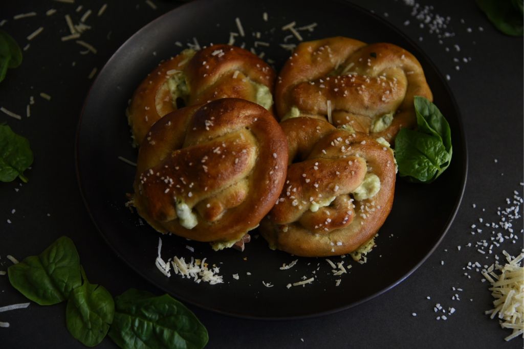 four pretzels on a plate with some salt, spinach and cheese around them