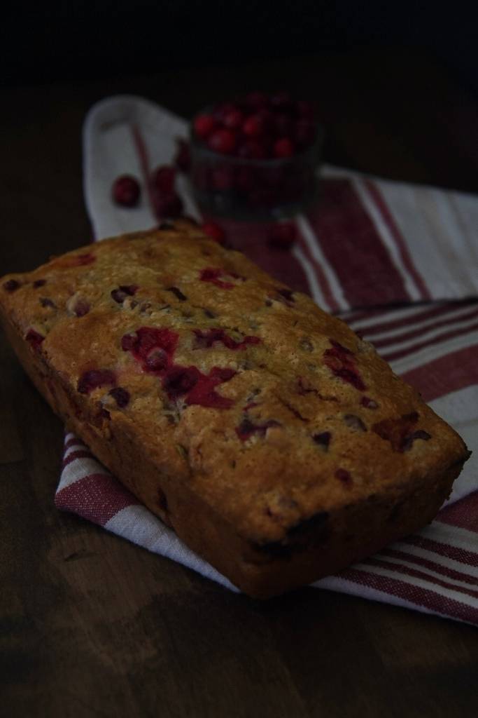 a loaf of cranberry orange bread sitting on a tea towel with a jar of cranberries behind it