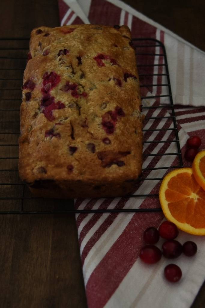 a loaf of bread on a cooling rack with a towel, orange slices and cranberry next to it