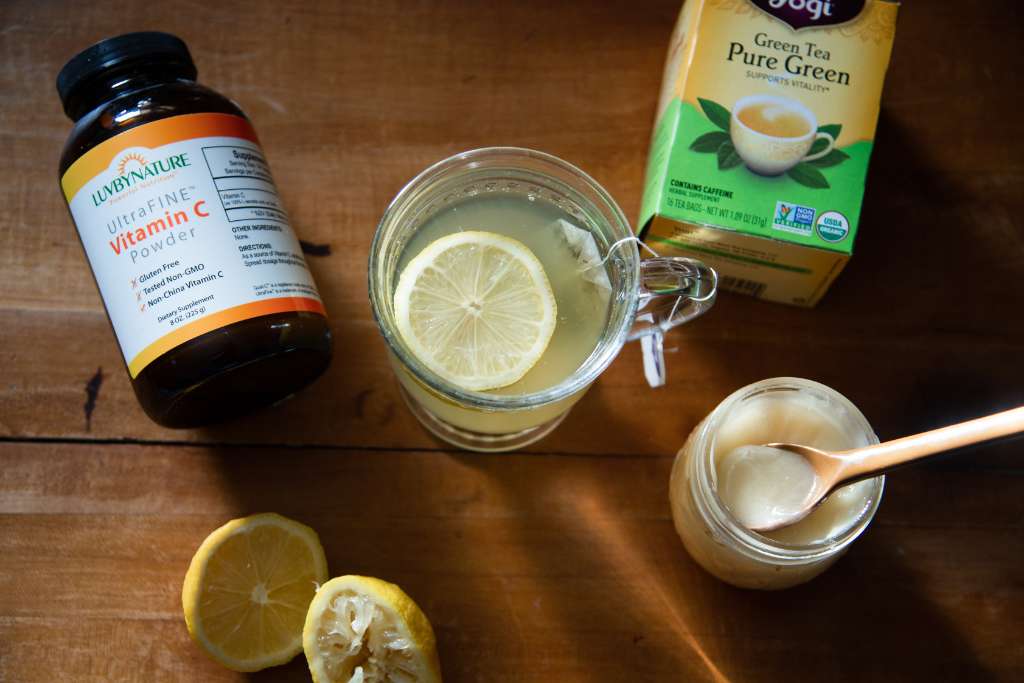 a cup of tea with a box of tea, lemons, honey, and vitamin C around it