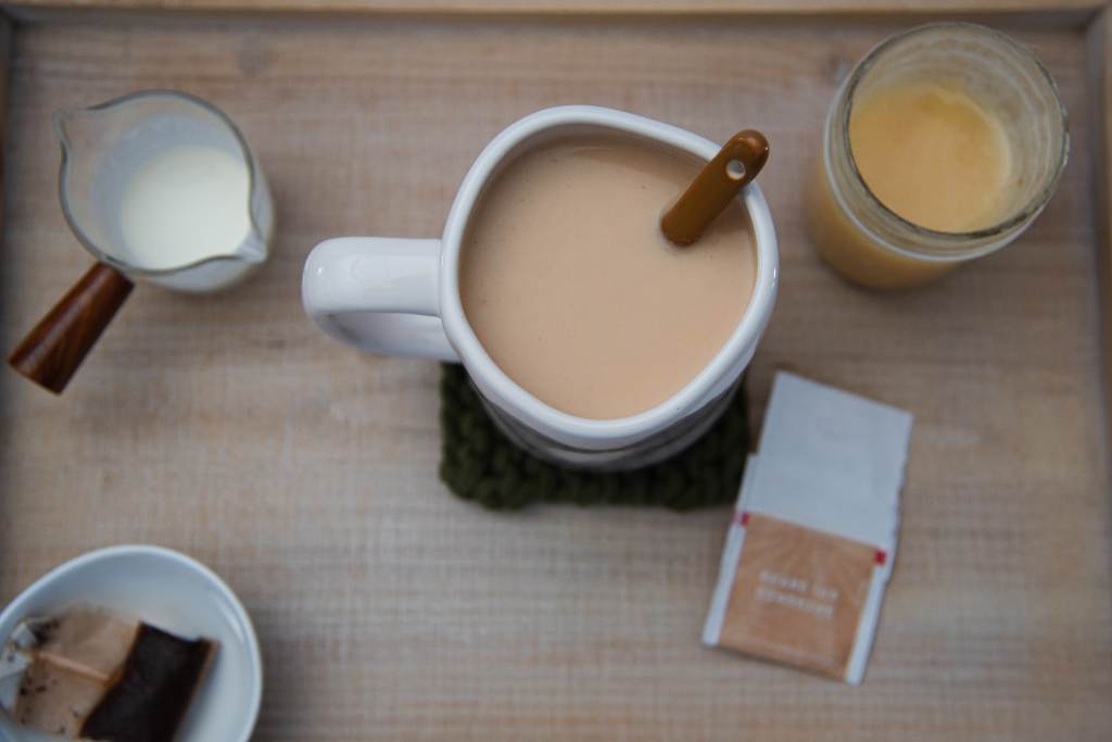 a finished black tea latte with a spoon in it. There is a tea bag, a jar of honey, and a dish of heavy cream next to it