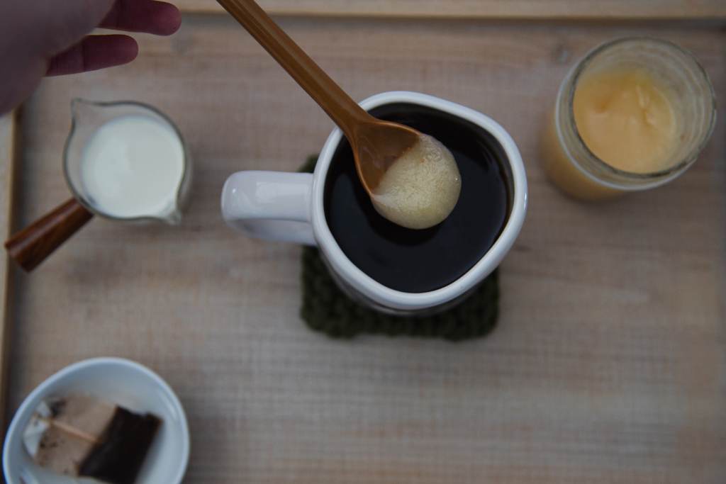 someone holding a spoonful of honey above a mug of black tea. There is a jar of honey, a dish of cream and tea bag around the mug