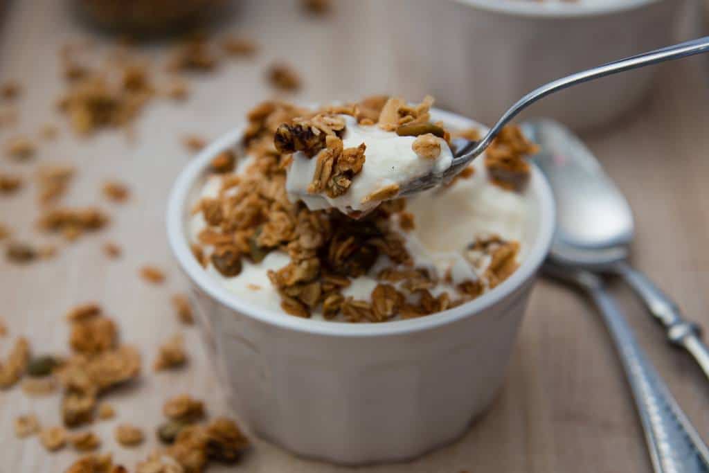 someone holding a spoon full of yogurt and granola over a dish of yogurt. there are some spoons and spilled granola in the back round