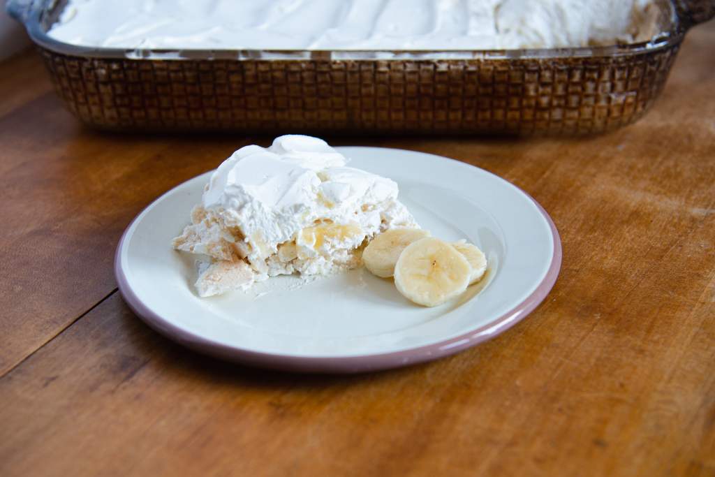 A piece of banana torte on a plate with three slices of banana next to it. And a pan torte behind it