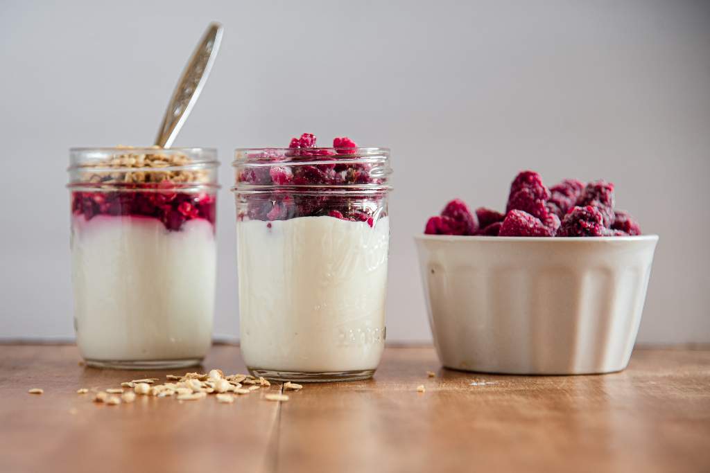 a jar of yogurt with frozen raspberries on it. There is a finished meal prep yogurt up with a spoon in it and a dish of frozen raspberries next to it