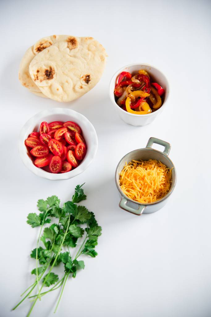 naan bread, caramelized onions and pepper,s halved cherry tomato, cheddar cheese and cilantro, in dishes on a white background