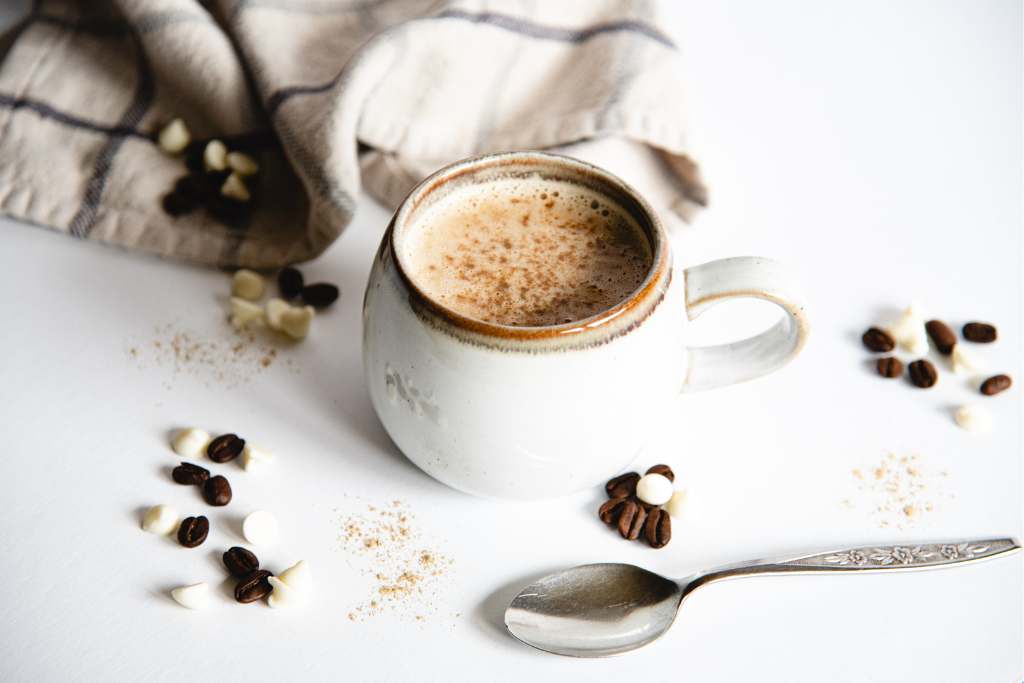 a finished latte in a mug with cardamom sprinkled on top of it. There are chocolate chips, coffees beans and cardamom sprinkled around it, a spoon is laying in front of the mug and there is a tea towel in the background  