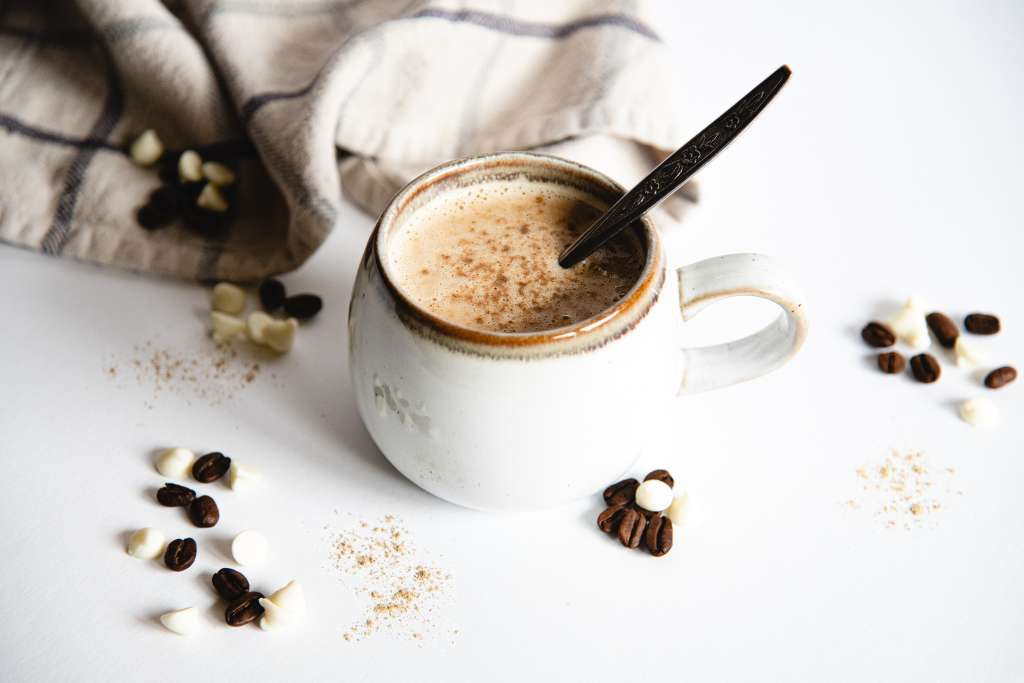 a mug of white chocolate espresso with a dusting on cardamom on it and a spoon in it. There are coffee beans, white chocolate chips and cardamom sprinkled around it, and a tea towel in the background 