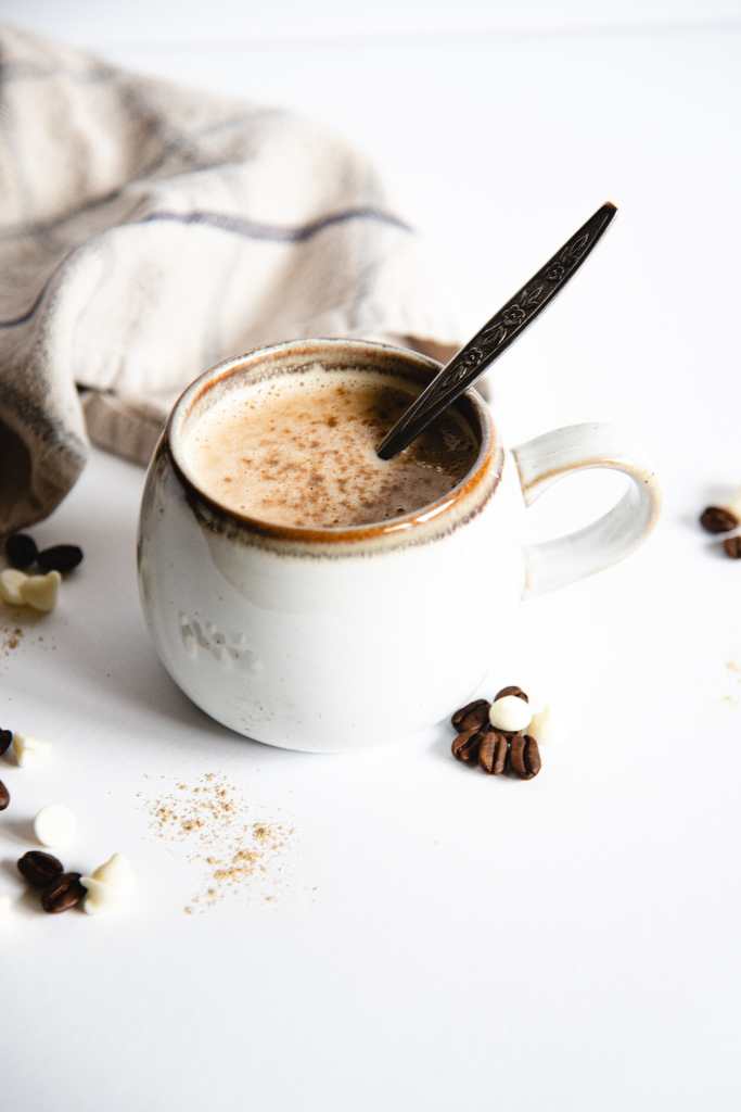 a mug of white chocolate espresso with a spoon in it. There are some coffee beans, white chocolate chips, and cardamom sprinkled around it, and you are see part of tea towel in the background 