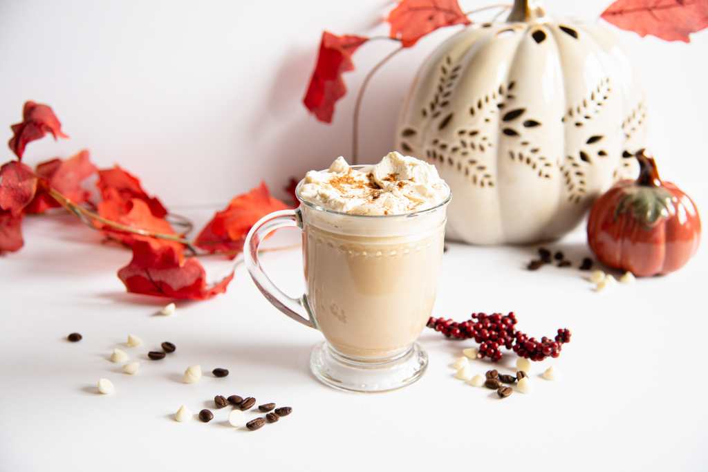 a finished latte in a clear glass cup, topped with whipped cream and cinnamon, there are fall decorations, chocolate chips and coffee beans around it 