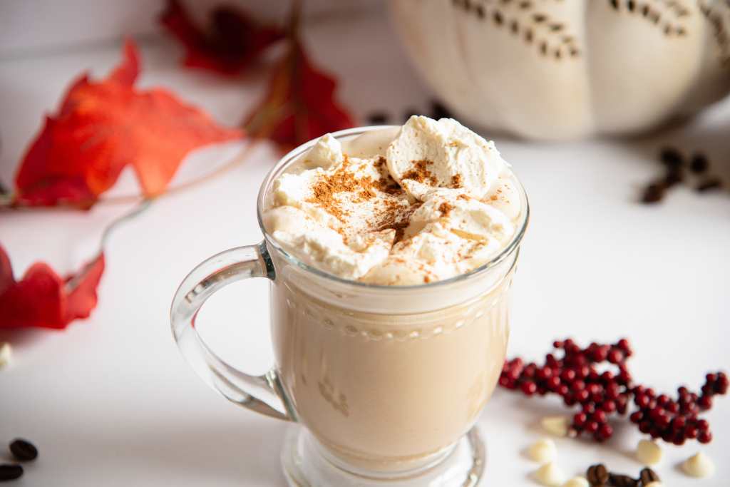 an overhead view of white chocolate pumpkin latte, with whipped cream and cinnamon on top, there are some fall decorations about it as well as some coffee beans and white chocolate chips