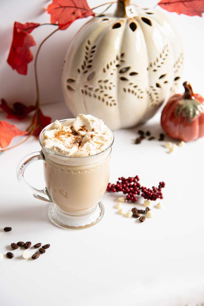 a finished latte topped with whipped cream and cinnamon, there are some white chocolate chips and coffee beans bu the cup and some fall decorations behind it