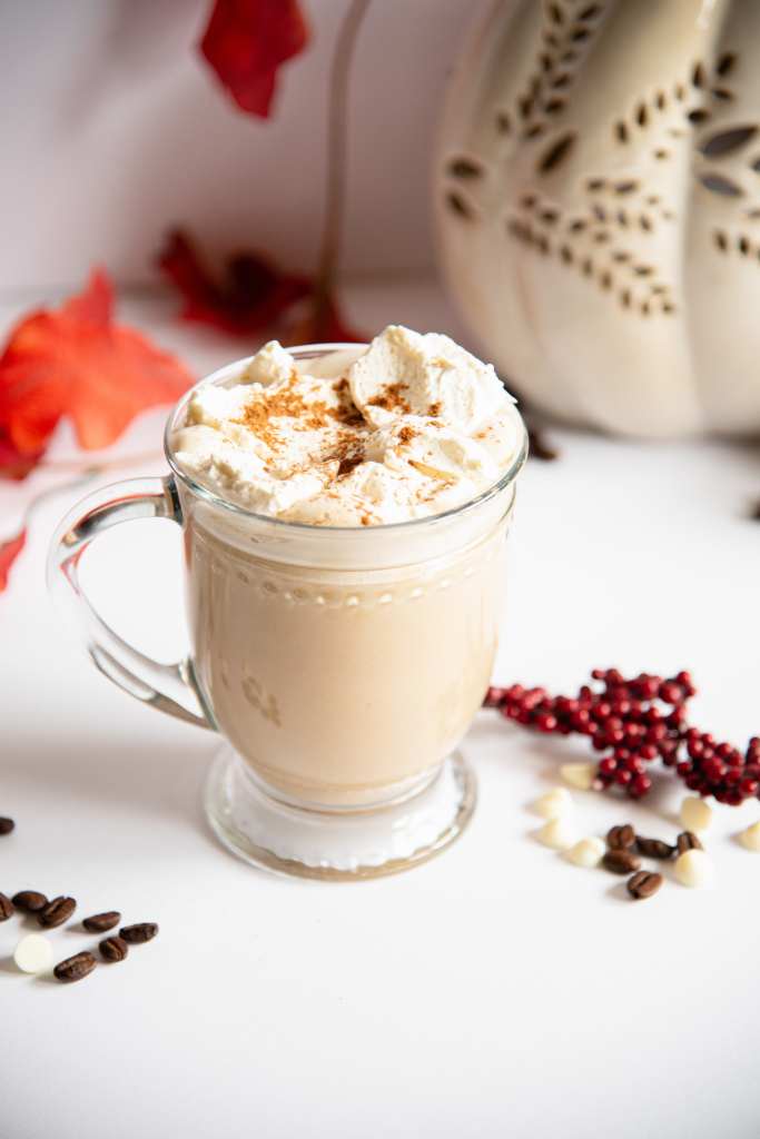 a white chocolate pumpkin latte topped with whipped cream and cinnamon, there are some white chocolate chips and coffee bean next to the cup and some fall decorations behind it