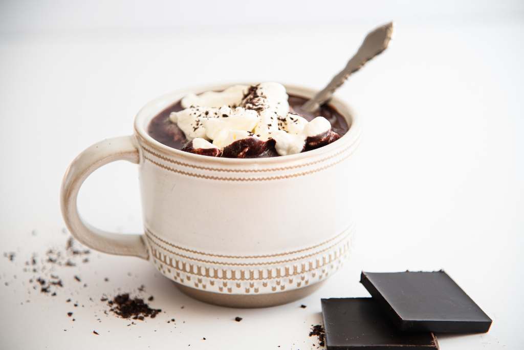 a side view of a mug of hot chocolate with whipped cream and a spoon in it, there are two pieces of chocolate next to the mug