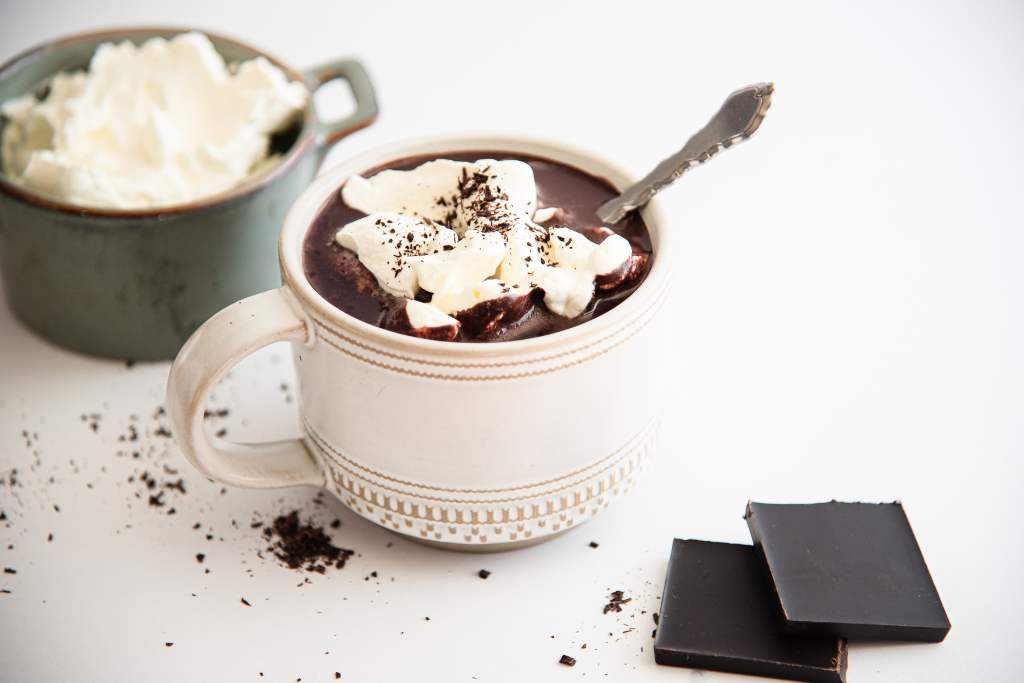 a mug of hot chocolate with whipped cream on top and a spoon in it. There is a dish of whipped cream and two pieces of chocolate next to the mug 
