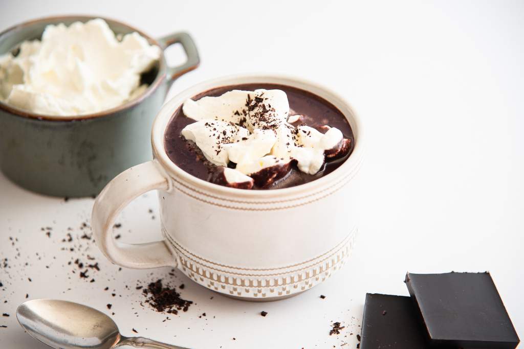a white mug of hot chocolate with whipped cream on top, there are two pieces of chocolate, a dish of whipped,c ream, and a spoon, next to the mug