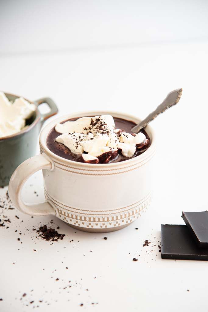 a mug of hot chocolate with whipped cream on top and a spoon in it, there are some chocolate pieces and a dish of whipped cream near the mug