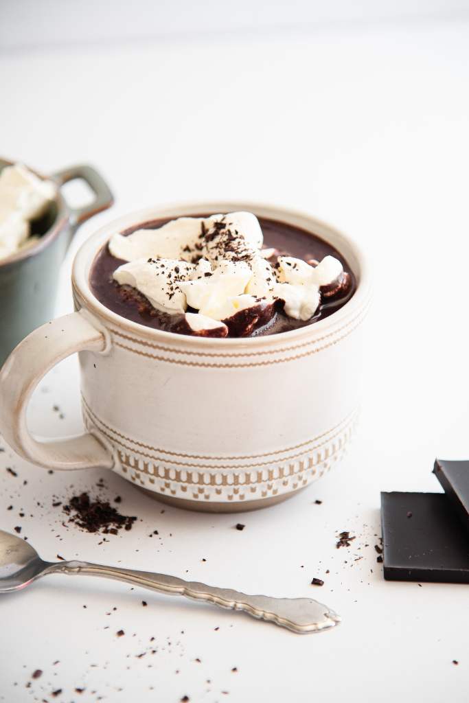 a close up of a mug of hot chocolate with whipped cream and chocolate shavings on top. You can see part of soon, a dish, and some chocolate pieces in the photo as well