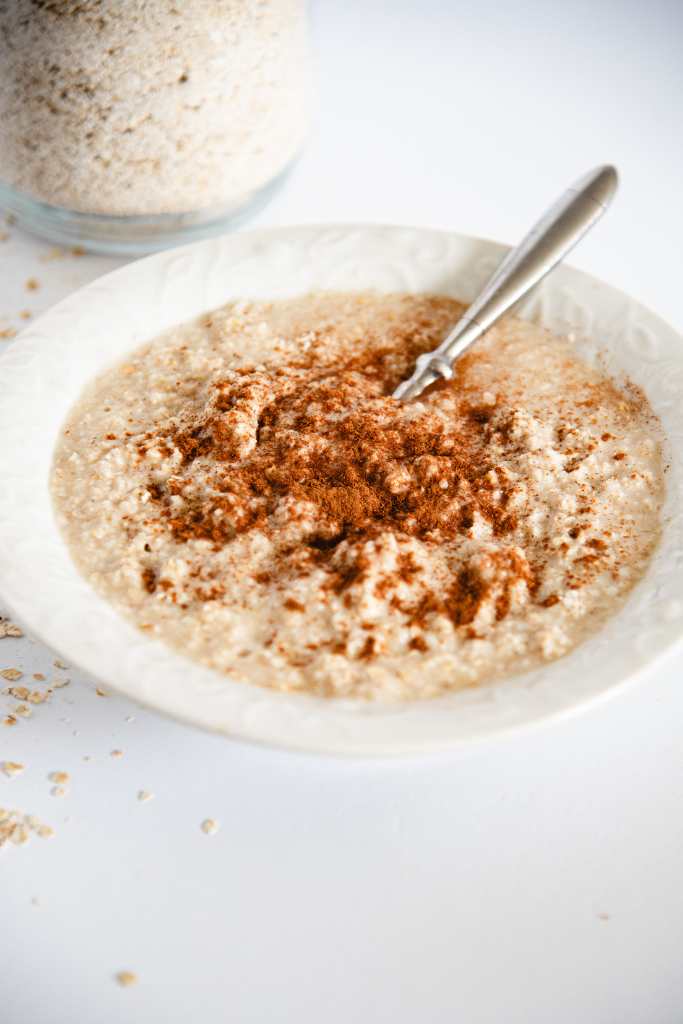 a bowl of cooked oatmeal with a spoon in it and cinnamon on top of it. You can see part of the container of oatmeal behind it