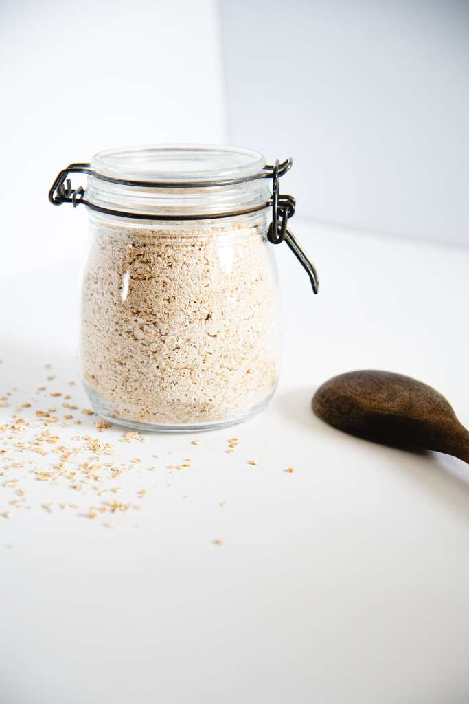 a closed glass container of instant oatmeal with a wooden spoon and some spilled oats by it