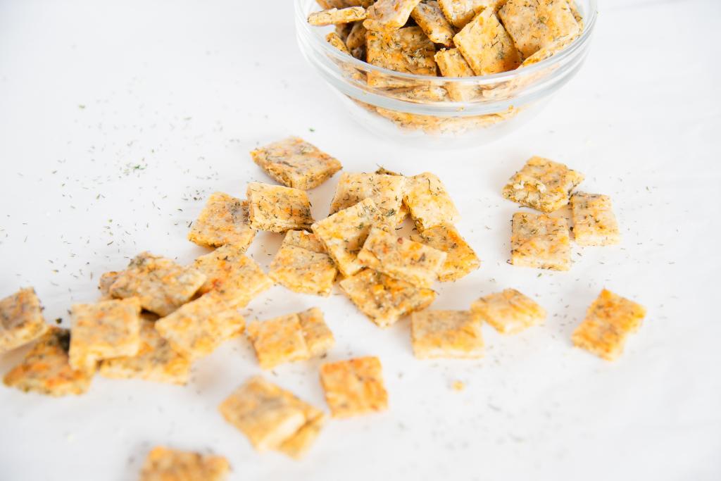 crackers and dill weed spilled on a table, you can see part of a glass bowl of crackers behind them