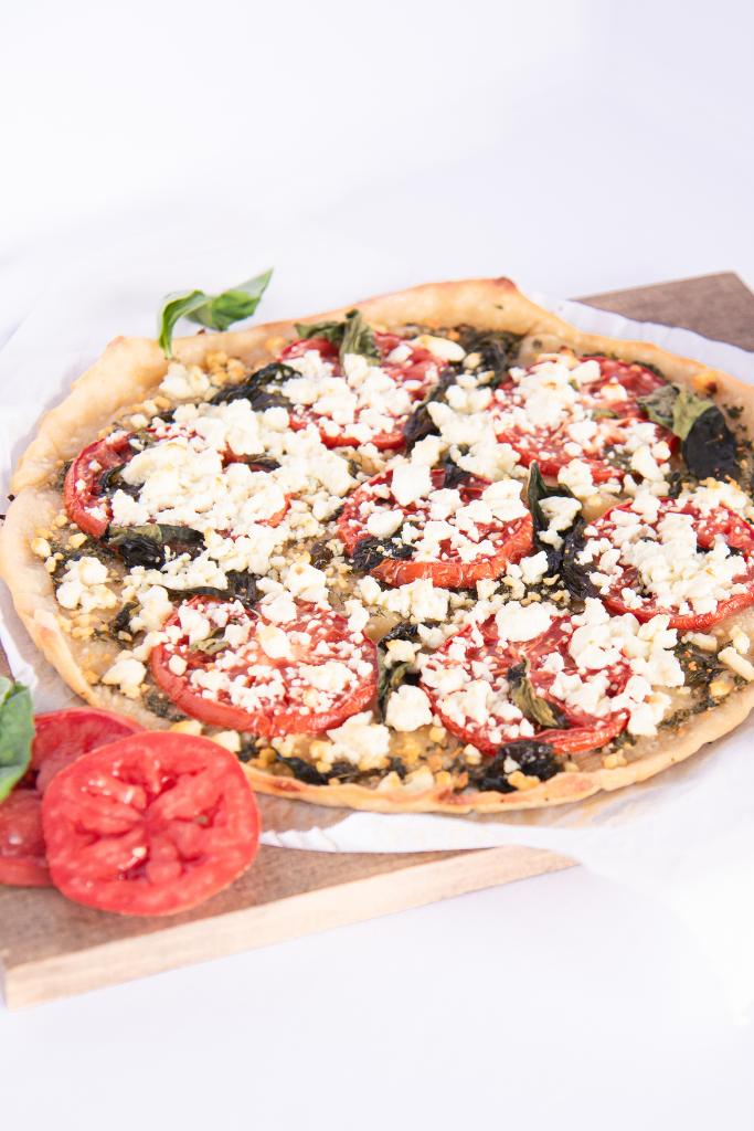 a feta and basil pizza on a wooden cutting board, with fresh tomato slices and basil leaves around it 
