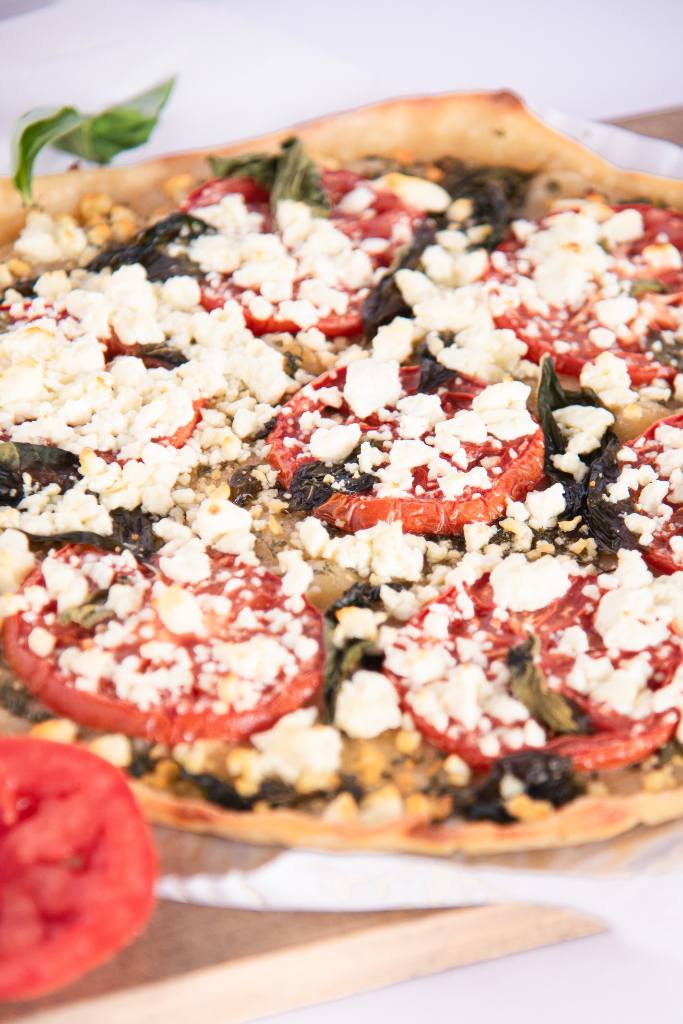 a feta and basil pizza on a wooden cutting board, there are some fresh tomato slices next to it as well